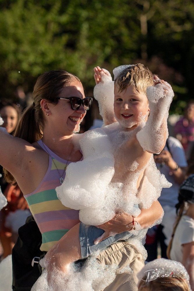 A woman wearing sunglasses and a colorful striped tank top is holding a young boy covered in soap suds, smiling and enjoying a bubble bath event outdoors in a park with people and green trees in the background.