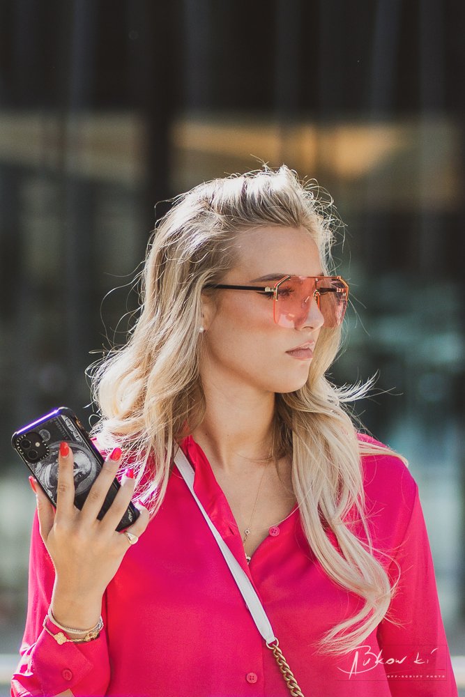 A young woman with long blonde hair wearing pink sunglasses and a vibrant pink blouse, holding a smartphone in her right hand, standing outdoors with a blurred cityscape background.