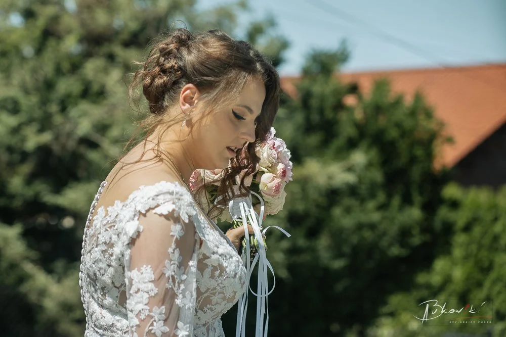 A bride in a lace wedding dress holding a bouquet of flowers with pink and white blooms, standing outdoors with greenery and a red rooftop in the background.