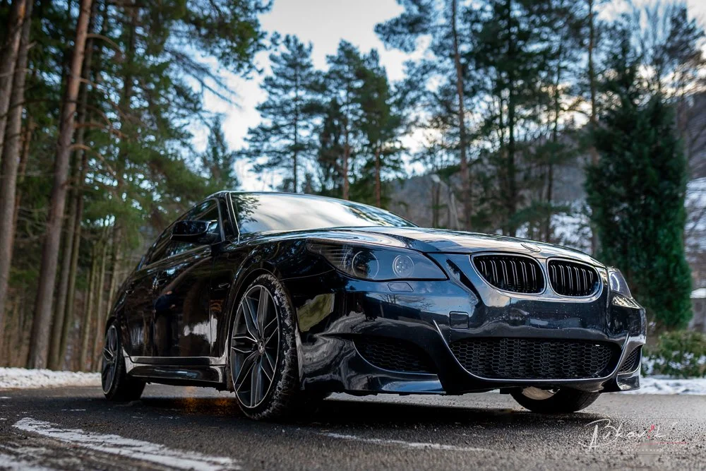 Black BMW car parked on a paved road surrounded by tall trees and snow in the background.