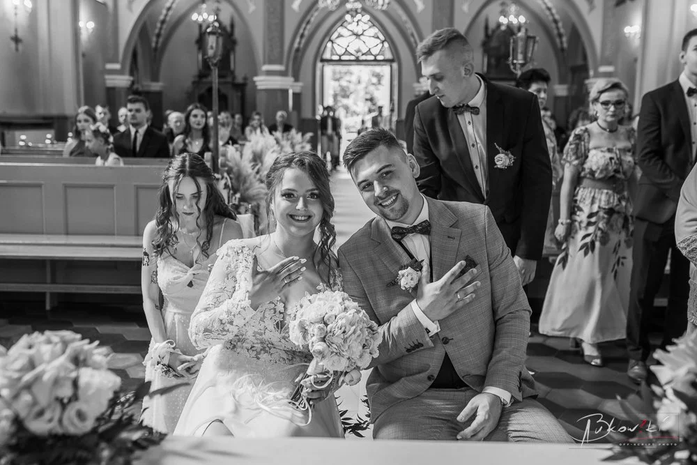 Black and white photo of a wedding ceremony showing a smiling bride and groom, with the bride showing her wedding ring, surrounded by guests in a church or chapel.