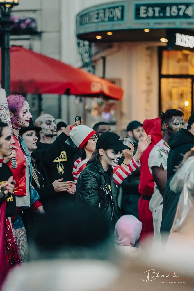 People dressed in Halloween costumes, including skeletons and clowns, gathered in a street with a storefront and a red awning.