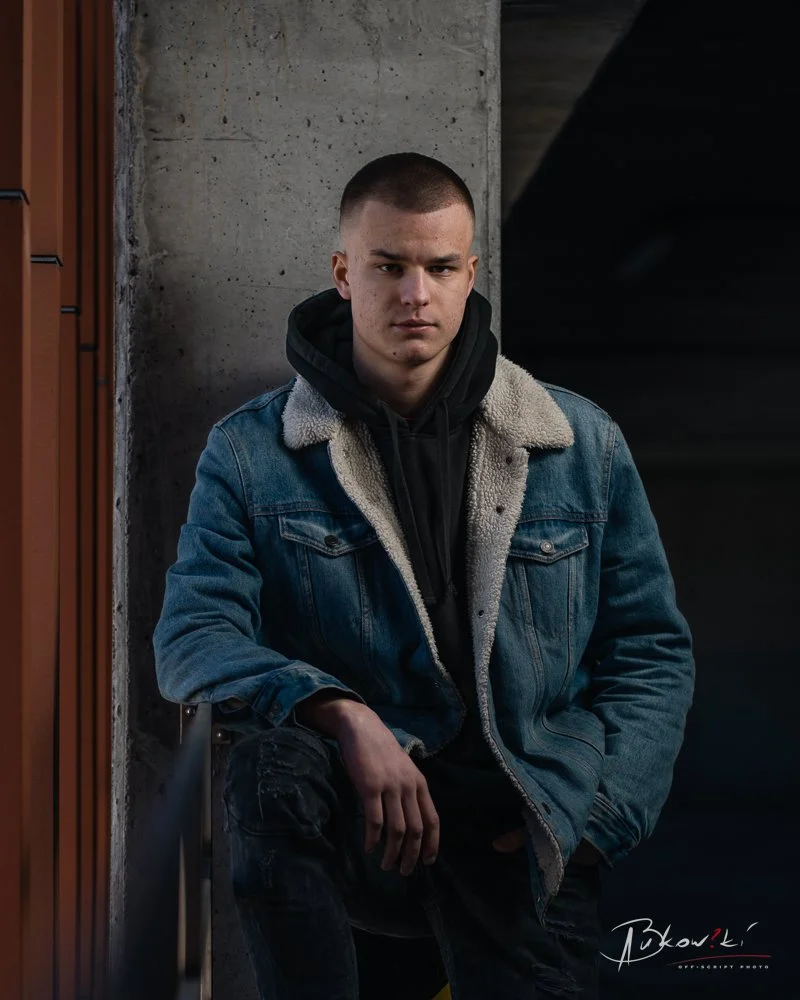Young man with short hair wearing a denim jacket and black hoodie, sitting against a concrete wall in a dimly lit, urban environment.