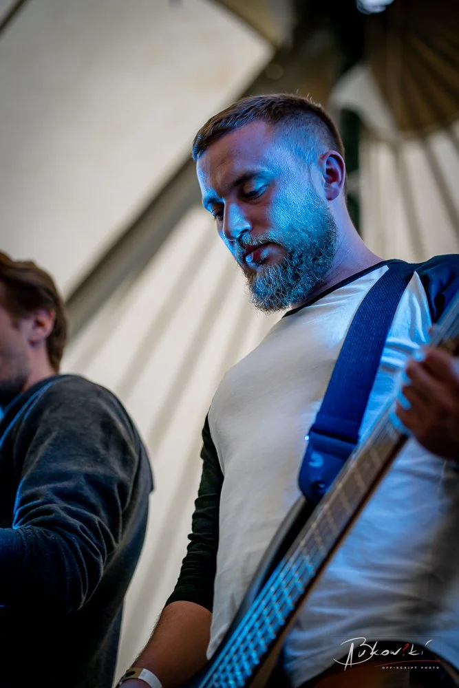 Man with a beard playing an electric guitar during a performance, with another person partially visible beside him. Blue lighting highlights the scene.