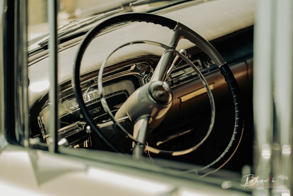 Close-up of the interior of a vintage car, showing the steering wheel and dashboard with the speedometer, viewed through the driver's side window.