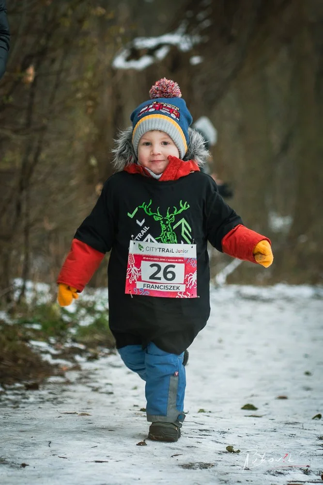 Young boy wearing winter clothes, running on snow-covered trail, participating in a race with bib number 26, smiling, in a wooded area.