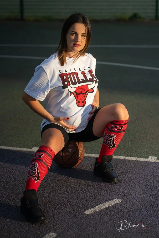 Young woman wearing a Chicago Bulls t-shirt and Milwaukee Bucks socks, sitting on a basketball on an outdoor court at sunset.