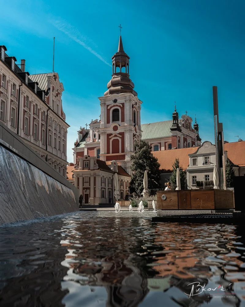 A historic building with a tall spire and ornate architecture, situated near a water feature with small fountains and umbrellas in an urban square under blue sky.