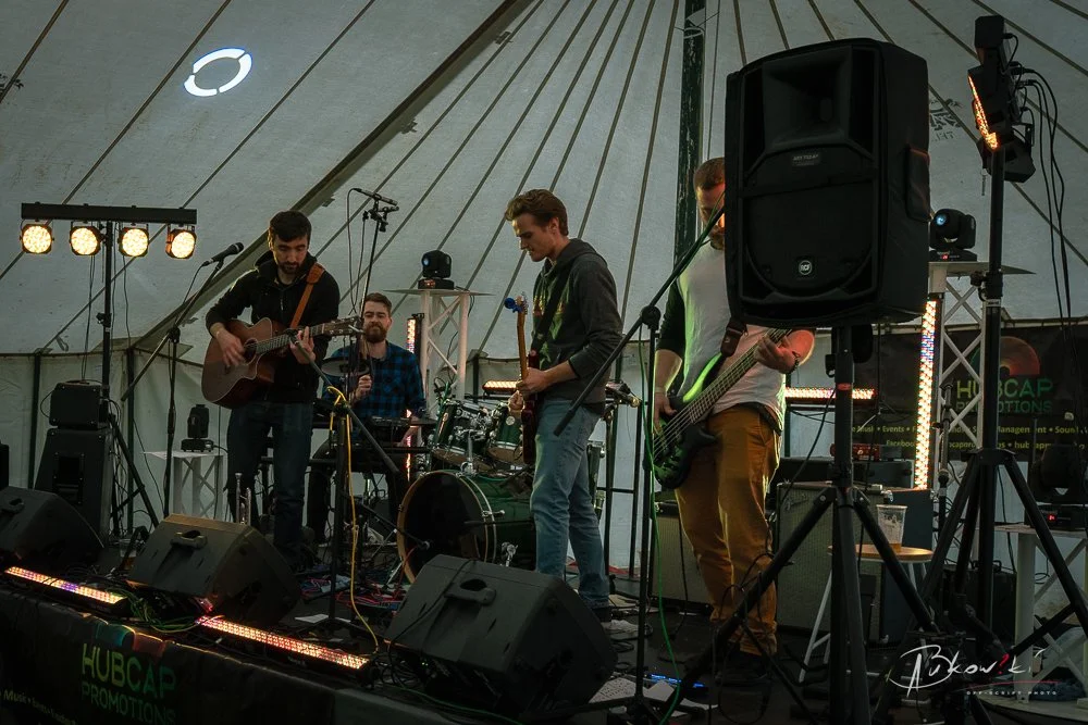 A band of four musicians performing on a stage inside a tent, with instruments including guitar, bass, drums, and microphone, surrounded by sound equipment and stage lights.