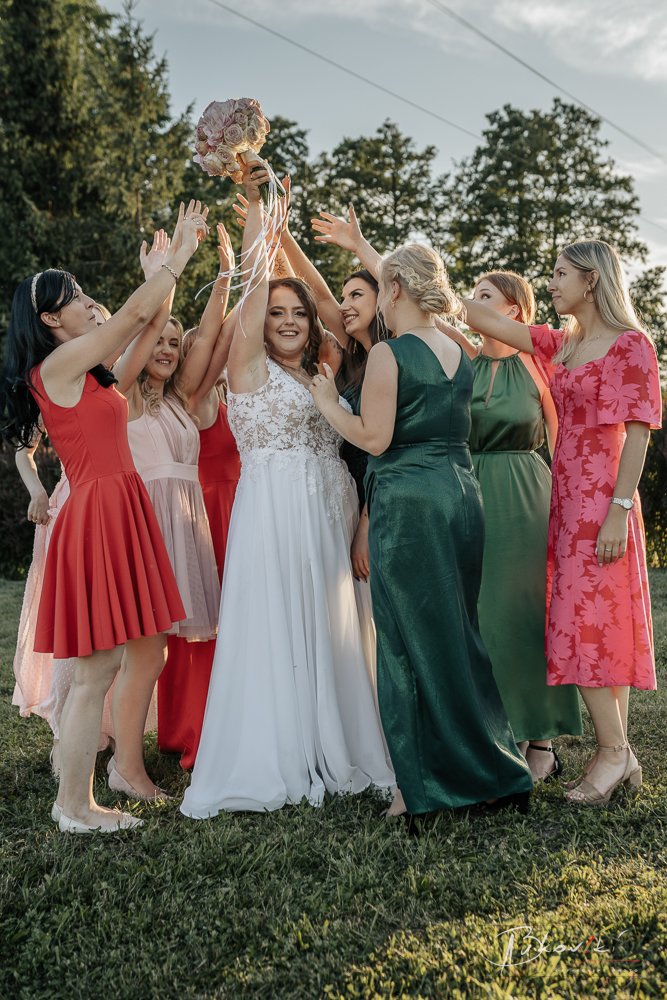 Group of women at a wedding celebration outdoors, with the bride holding a bouquet of flowers high, surrounded by friends in colorful dresses, cheering and celebrating.