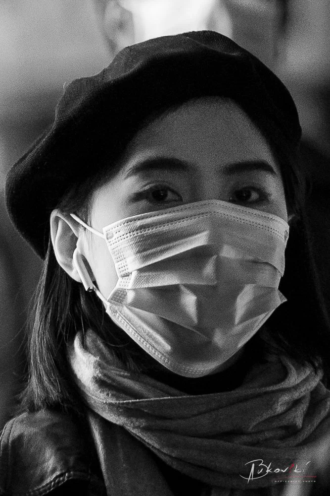 Close-up grayscale portrait of a woman wearing a face mask, scarf, and beret, with background blurred.