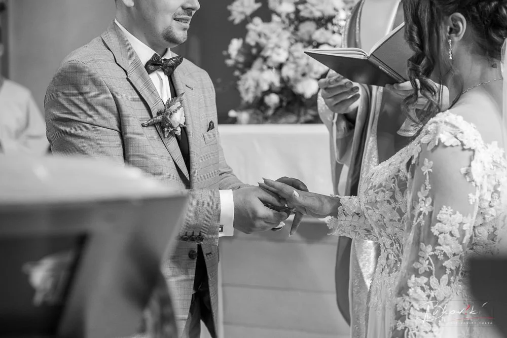 A bride and groom exchanging wedding rings during a ceremony, with a priest present, in black and white.