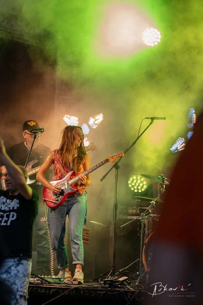 A young woman with long hair in casual clothes playing an electric guitar on stage with colorful stage lighting and fog, accompanied by other musicians.