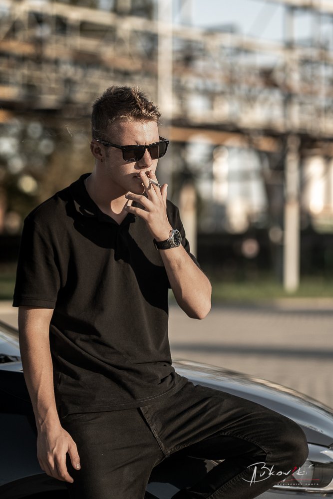 Young man wearing sunglasses, black shirt, and watch, smoking a cigarette while leaning against a car outdoors with a blurred background of buildings and power lines.