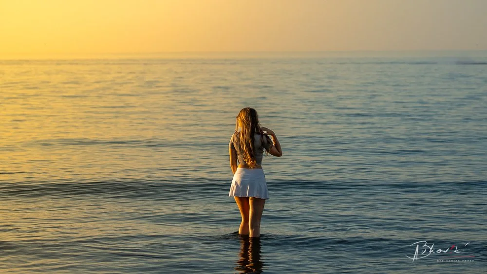 A woman in a gray top and white skirt stands in the ocean at sunset, facing away from the camera, with her long hair flowing down her back.