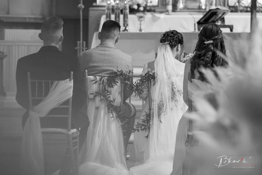 Four people sitting in chairs during a wedding ceremony, with the bride wearing a veil and the groom and others dressed in suits, decorated with white fabric and greenery in a bright indoor setting.