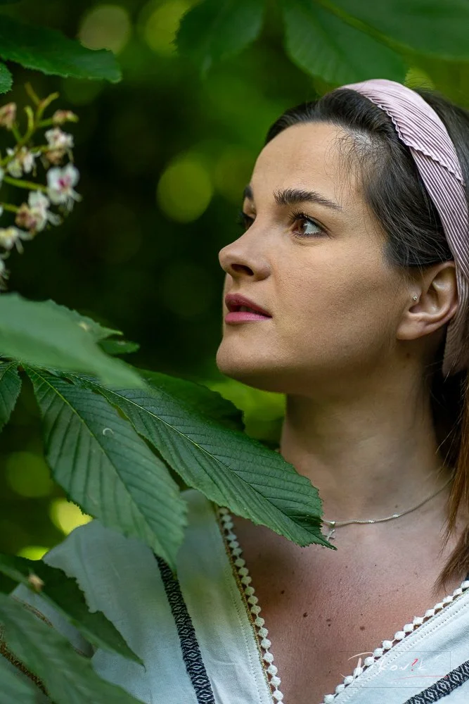 A woman with brown hair, wearing a pink headband and a white top, is outdoors among green leaves and trees, looking to her left.