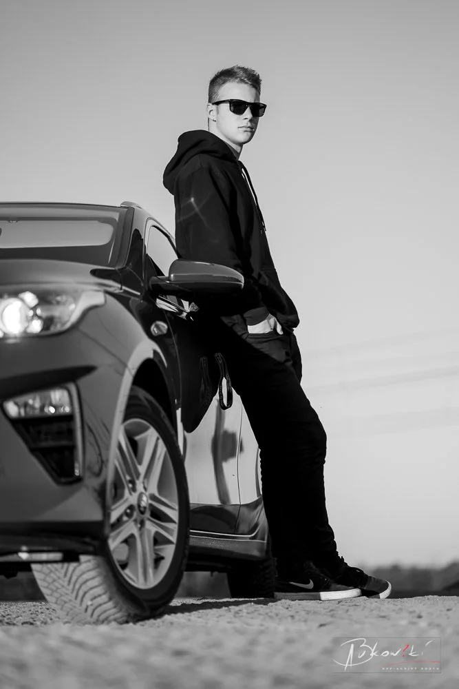 A young man wearing sunglasses and a hoodie leans against a car in an outdoor setting, with a clear sky in the background.