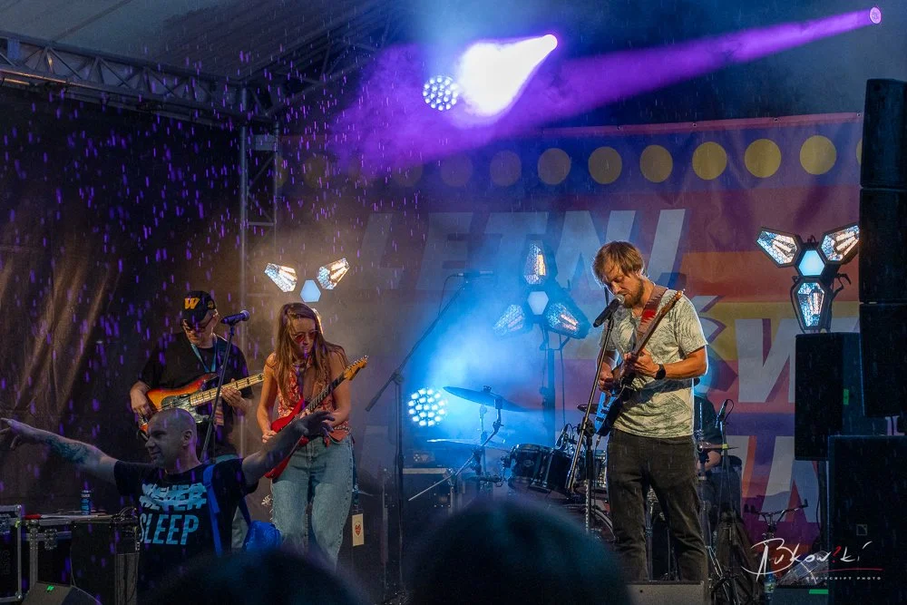 A band performing live on stage with colorful stage lighting and a fog effect, featuring a guitarist, bassist, and drummer.