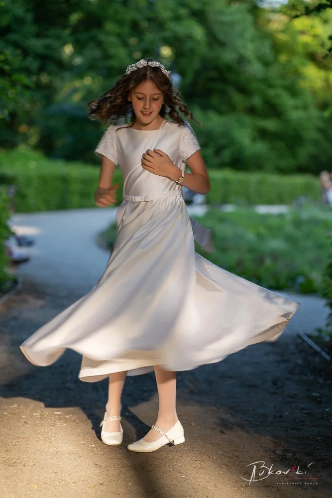 A young girl in a white dress and white shoes spinning and smiling outdoors on a dirt path surrounded by green trees.