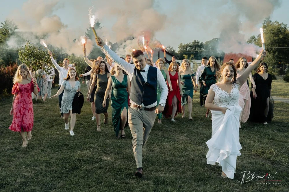 A wedding celebration outdoors with a bride and groom walking forward, holding sparklers. The bride is in a white wedding dress, smiling, and the groom is in a vest and tie. Behind them, friends and family are also holding sparklers and celebrating.
