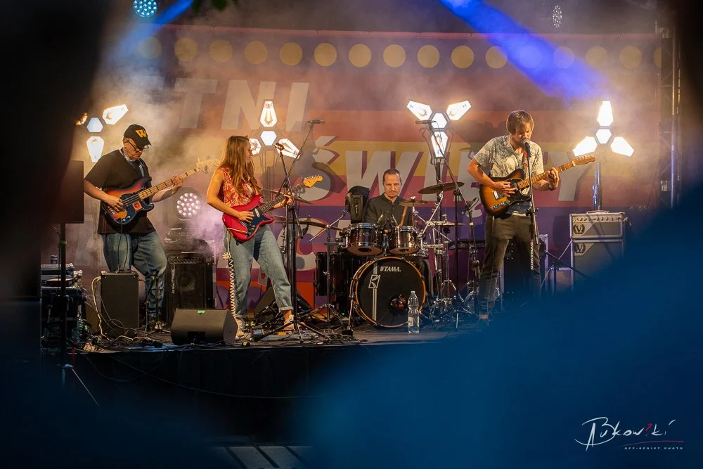 A live band performs on stage with four musicians playing guitars, drums, and bass amidst colorful lights and fog effects.