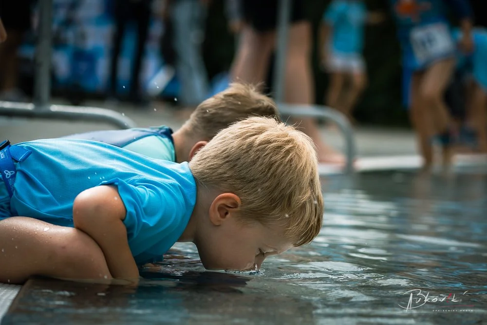 A young boy with blond hair in a blue shirt leans over the edge of a swimming pool, drinking or playing in the water, with other children in the background.