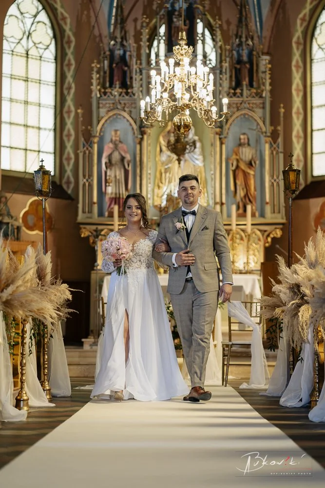 A bride and groom walking down the aisle inside a church, with the bride holding a bouquet of pink flowers and the groom dressed in a gray suit. The church features large stained glass windows, a chandelier, and religious statues.