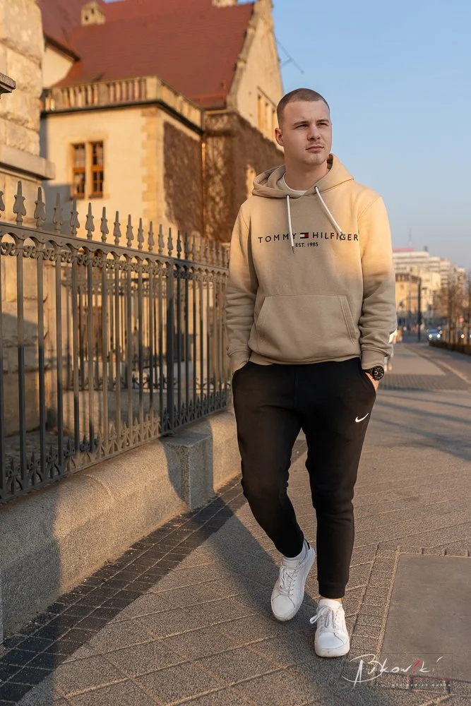 A young man standing on a sidewalk next to a black metal fence with his hands in the pockets of his black Nike sweatpants. He is wearing a beige Tommy Hilfiger hoodie, white sneakers, and a watch. The background features a historic building with a re