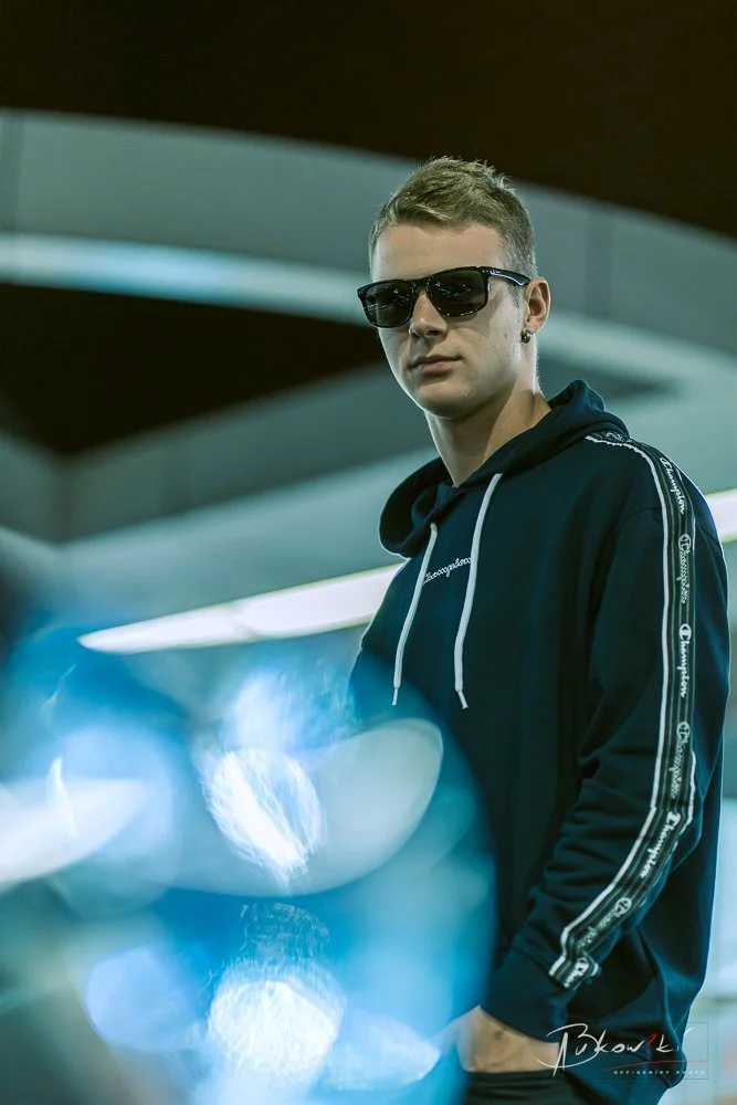Young man wearing sunglasses and a black hoodie with white accents, standing indoors near a blue-lit display.