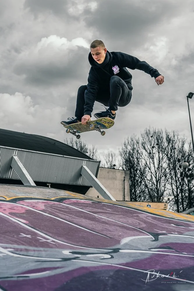 A young man wearing a black hoodie and black pants is skateboarding in mid-air over a ramp at an outdoor skate park on a cloudy day.