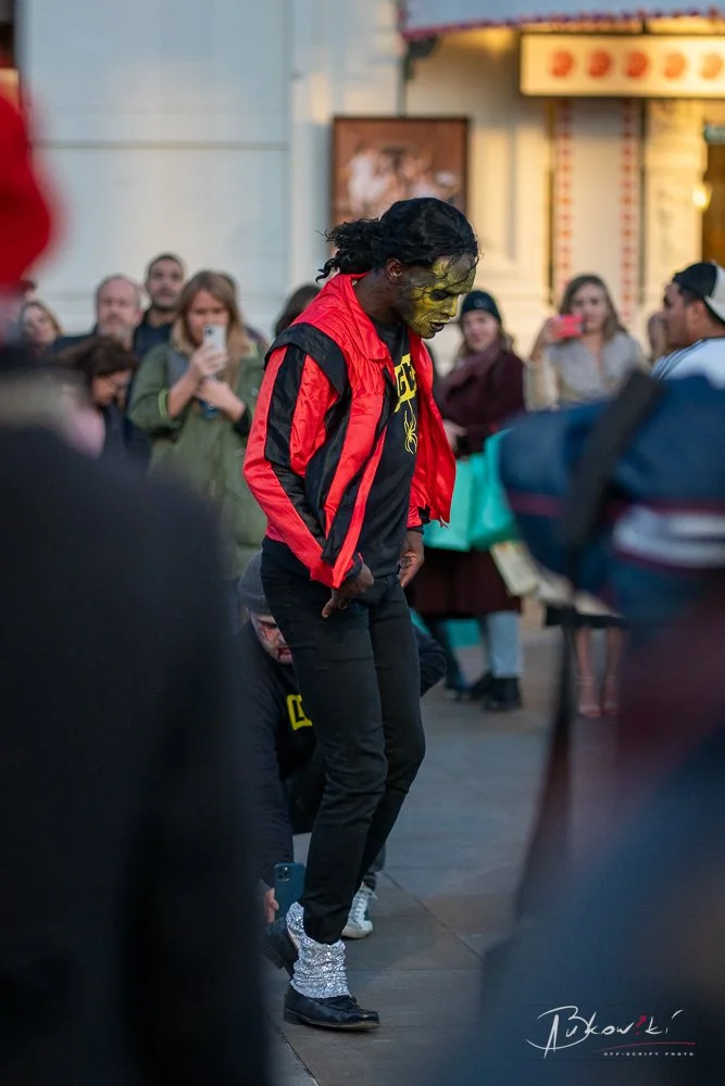 A performer with yellow face paint, long hair, and sunglasses, dressed in a red and black jacket, dancing in front of a crowd at an outdoor event.