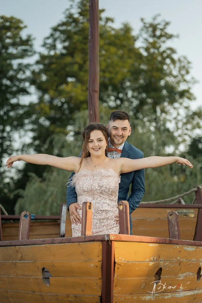 A smiling woman with arms outstretched and a smiling man in a blue suit standing behind her on a wooden boat, with trees in the background.
