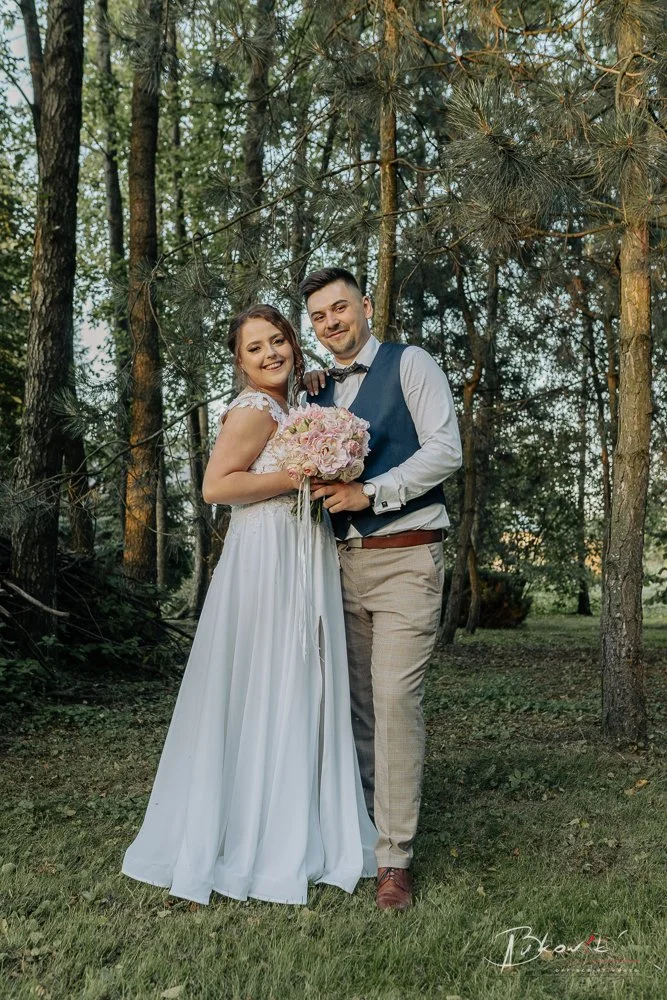 Happy couple standing in a forest, the woman in a white wedding dress holding a bouquet of pink flowers, the man in a blue vest and beige pants, both smiling at the camera.
