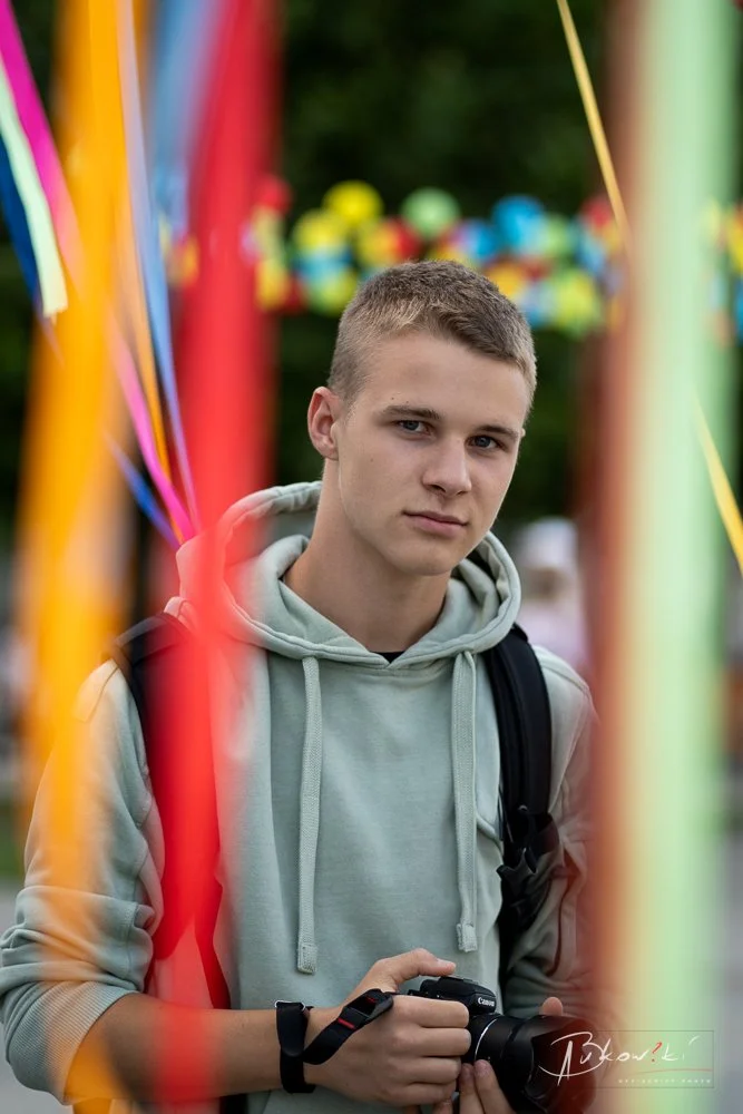 A young man with short hair wearing a light gray hoodie, carrying a black backpack, holding a camera, standing outdoors at a colorful amusement park or fairground with blurred bright yellow, red, orange, and green rides or decorations in the backgrou