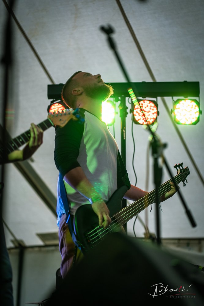A musician with a beard and short hair playing an electric bass guitar on stage, with colorful stage lights behind him, and another guitarist partially visible to the side.