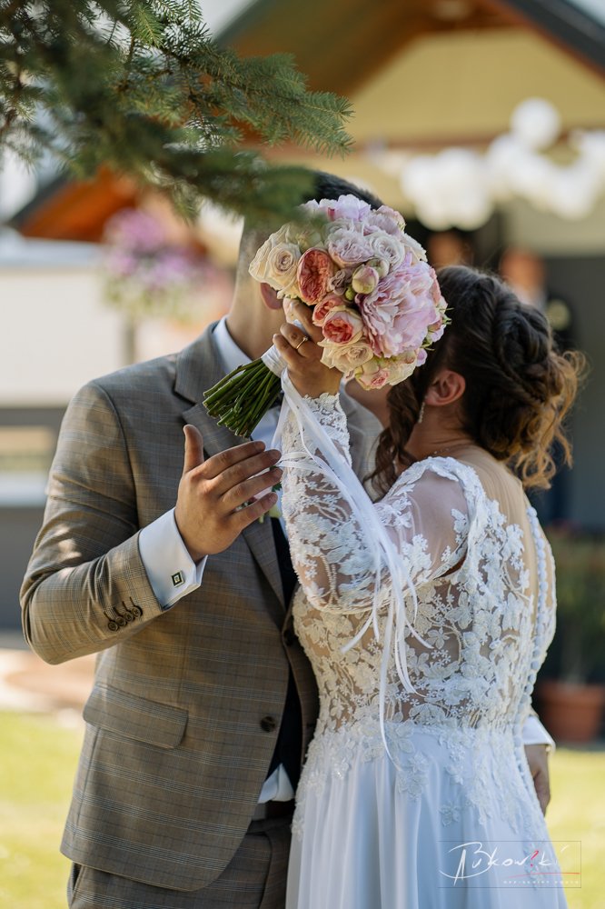 A bride and groom share a kiss behind a bouquet in an outdoor wedding setting, with blurred background and greenery.