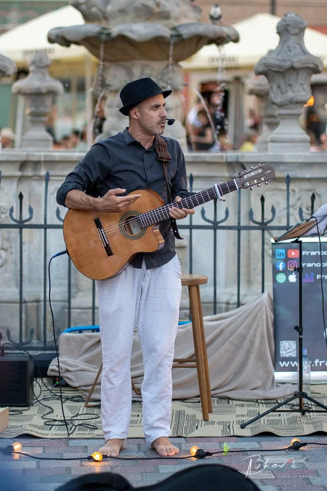A man with a black hat and a microphone headset plays an acoustic guitar while performing barefoot outdoors near a fountain with stone sculptures. There is a stool and an electronic sign with social media icons beside him.