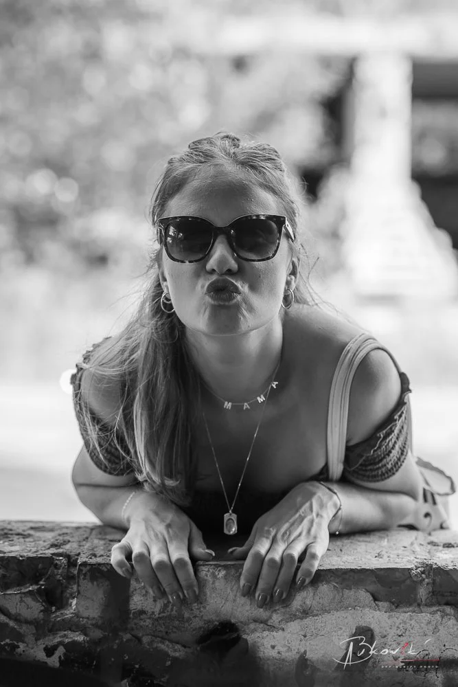 A woman with long hair, wearing sunglasses, making a kissing face, leaning on a stone surface, outdoors in a sunny area.