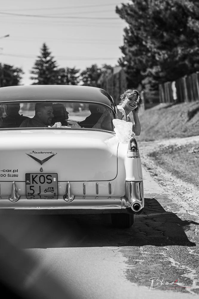 Black and white photo of a happy woman sticking her head out of a vintage car with a wedding dress, while three people sit inside the car in conversation.
