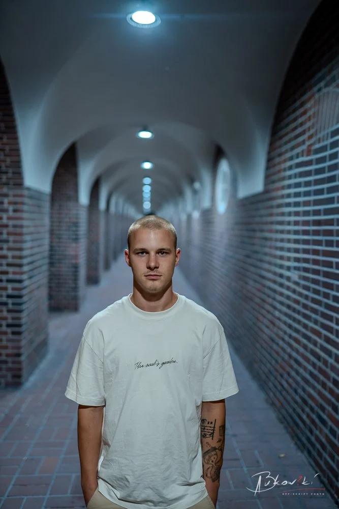 A young man with a buzz cut hairstyle standing in a dimly lit brick corridor with brick walls and arched ceiling.