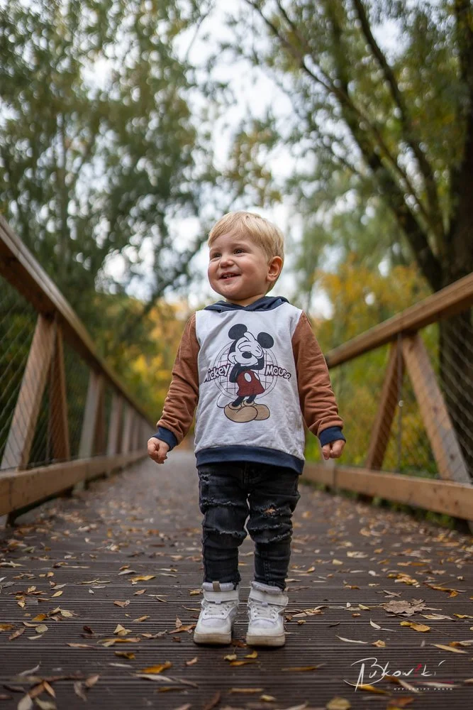 A young child with blonde hair smiling and walking on a wooden bridge surrounded by green trees and fallen leaves.
