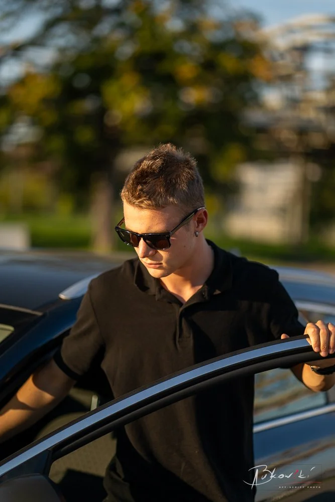 Young man with short brown hair wearing sunglasses and a black polo shirt stepping out of a car during sunset with a blurred outdoor background.