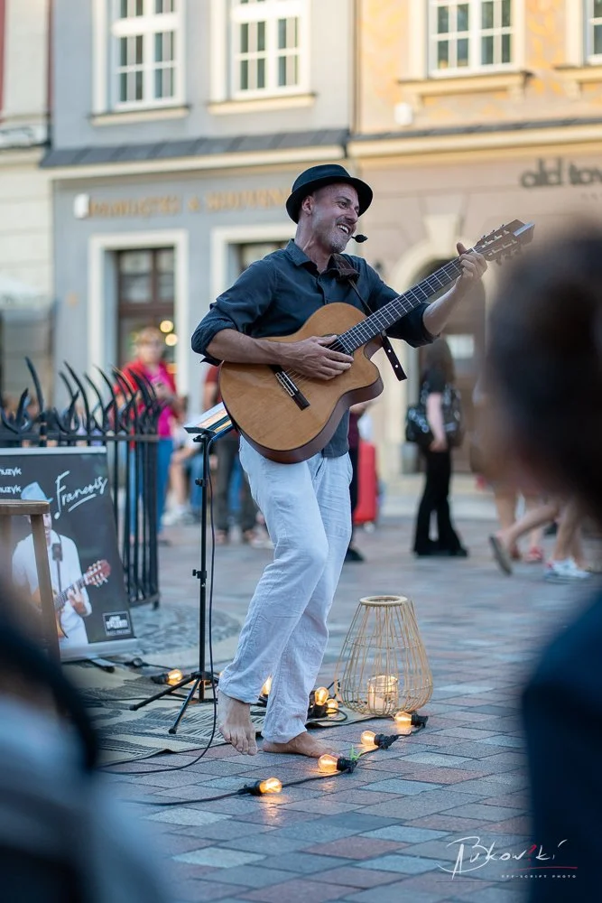 A street musician with a hat and headset playing an acoustic guitar barefoot on a city pedestrian street, surrounded by string lights and lanterns, with blurred passersby in the background.