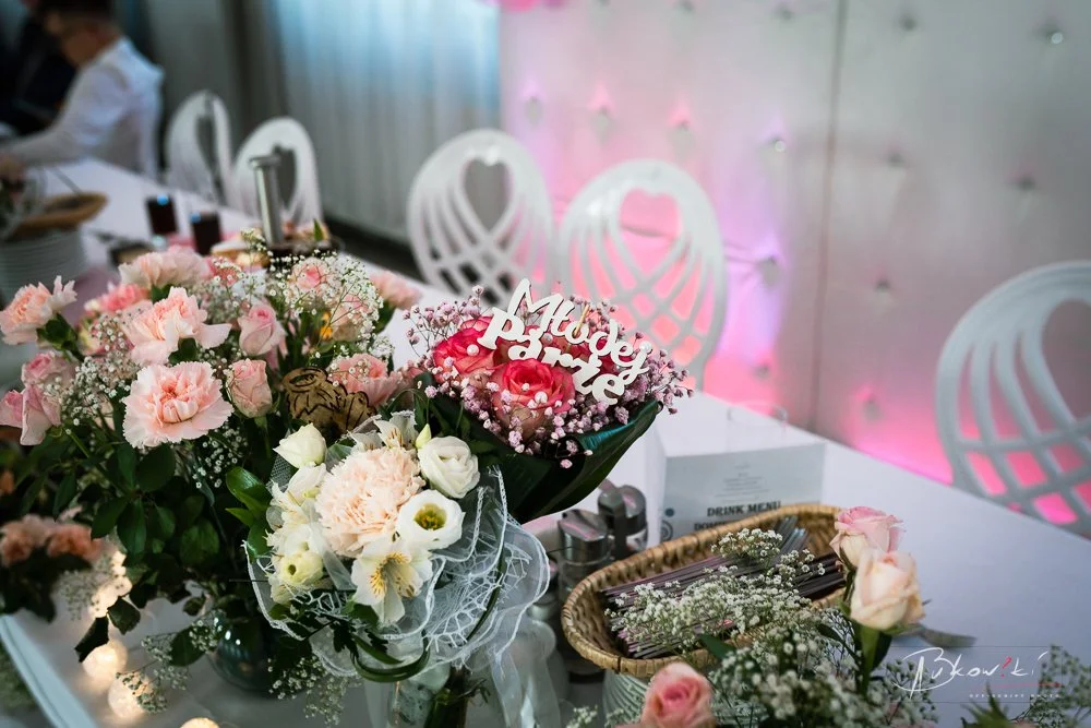 Table decorated with pink and white flowers, including roses and baby's breath, with a sign that says "Model of the Bride," in a decorated banquet hall.