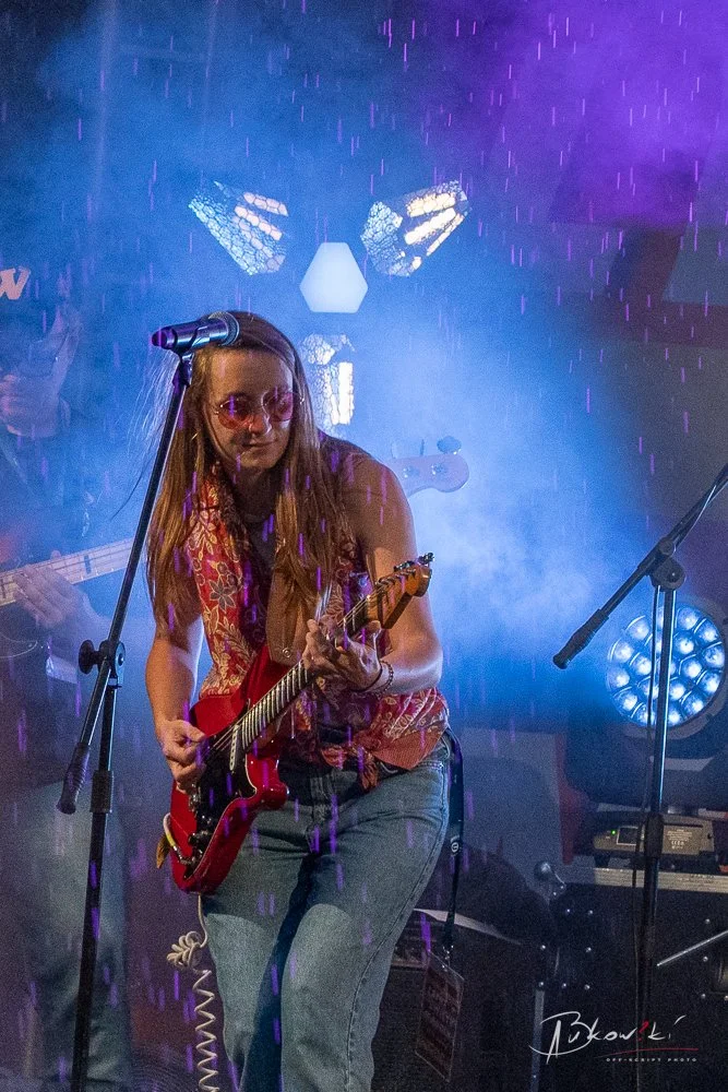 A woman playing an electric guitar on stage with purple and blue lighting, wearing sunglasses and a patterned top.