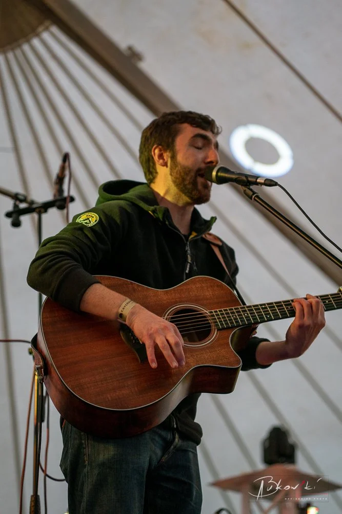 A man with dark hair and a beard sings into a microphone while playing an acoustic guitar on stage under a tent.