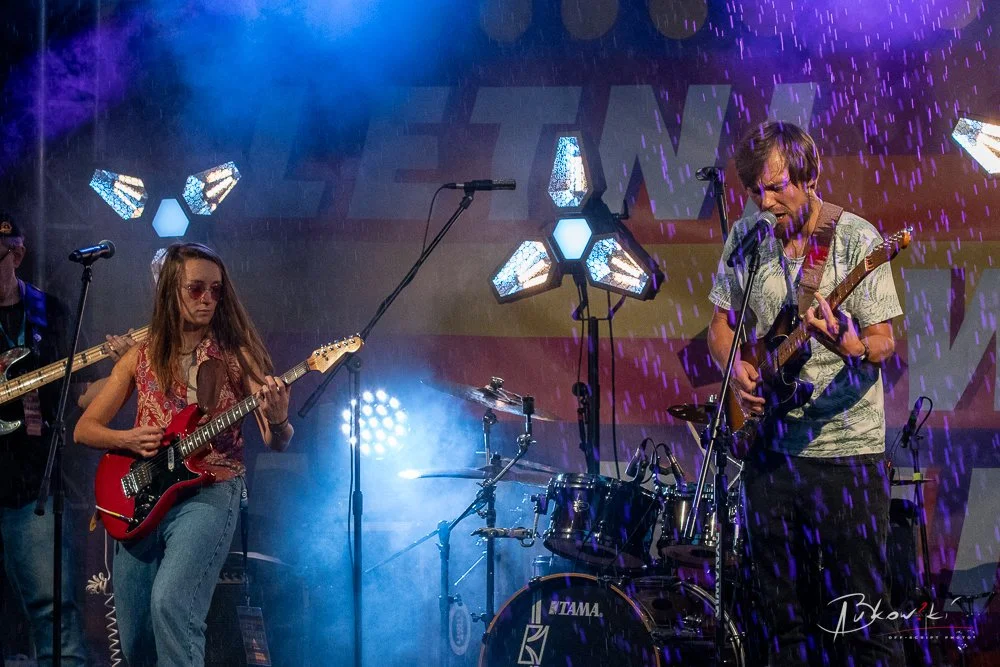 Musicians performing on stage with guitars and drums, colorful stage lights and rain effects.