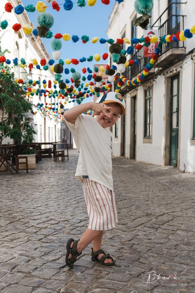 A young boy standing on a cobblestone street decorated with colorful paper lanterns hanging above, making a playful gesture with his hand near his eye and smiling.