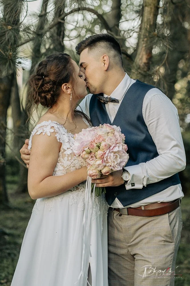 A bride and groom sharing a kiss outdoors, holding a bouquet of pink and white flowers, with trees in the background.